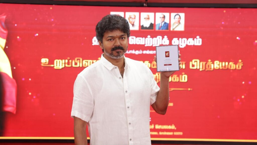 Actor Vijay launches TVK mobile app at an official party event, wearing a white shirt and holding a tablet against a red party backdrop with political leaders’ portraits.